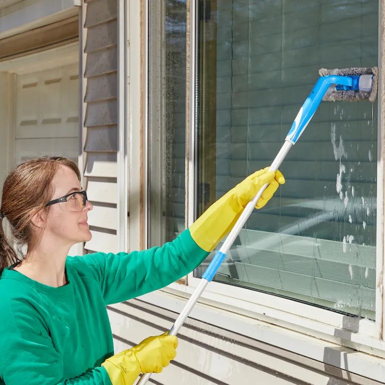 A woman in a green shirt and yellow gloves diligently cleans a window, illustrating a window cleaning service in action.